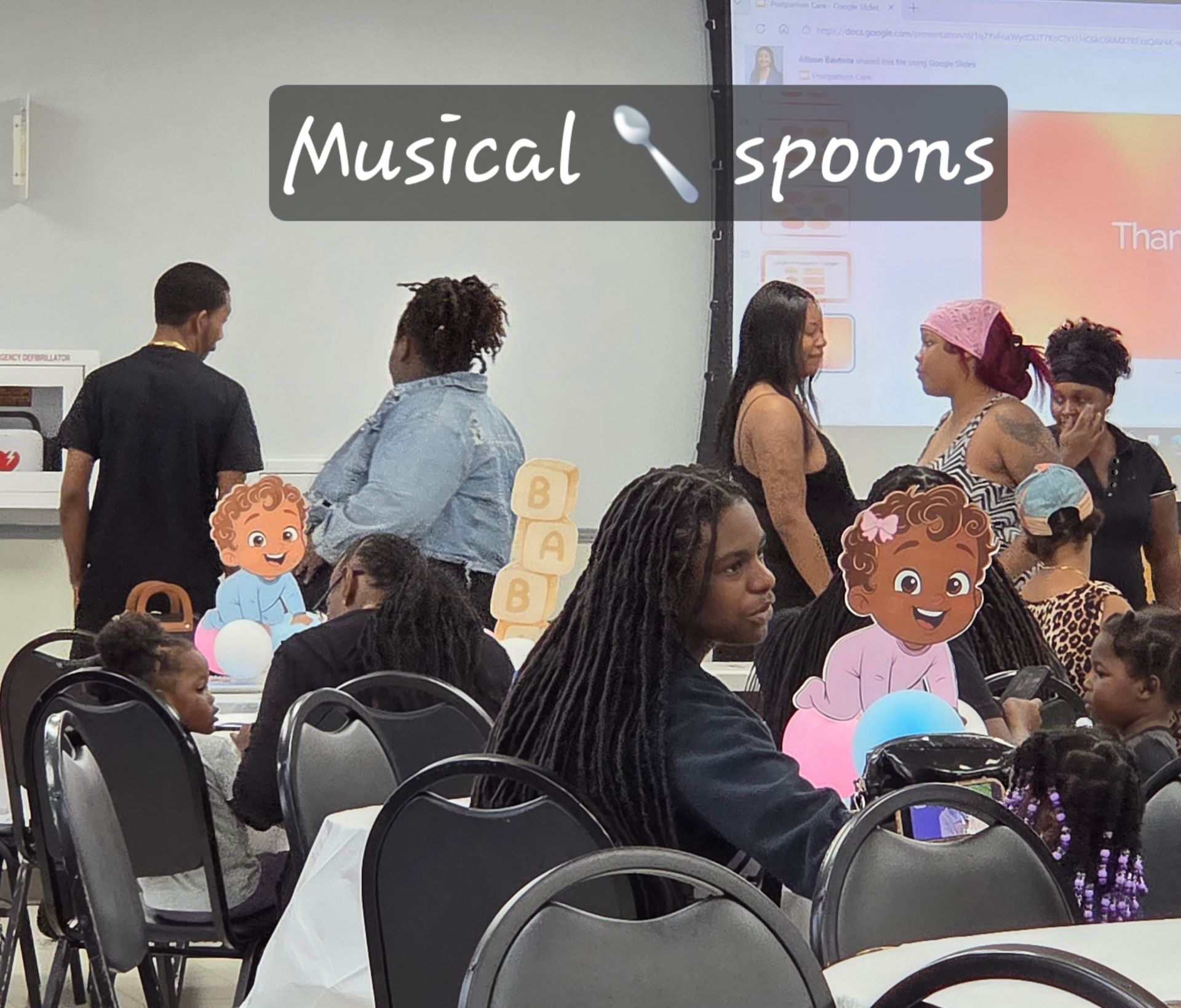 People gathered in a classroom for a “Musical spoons” activity, with chairs and children’s cutout faces visible.