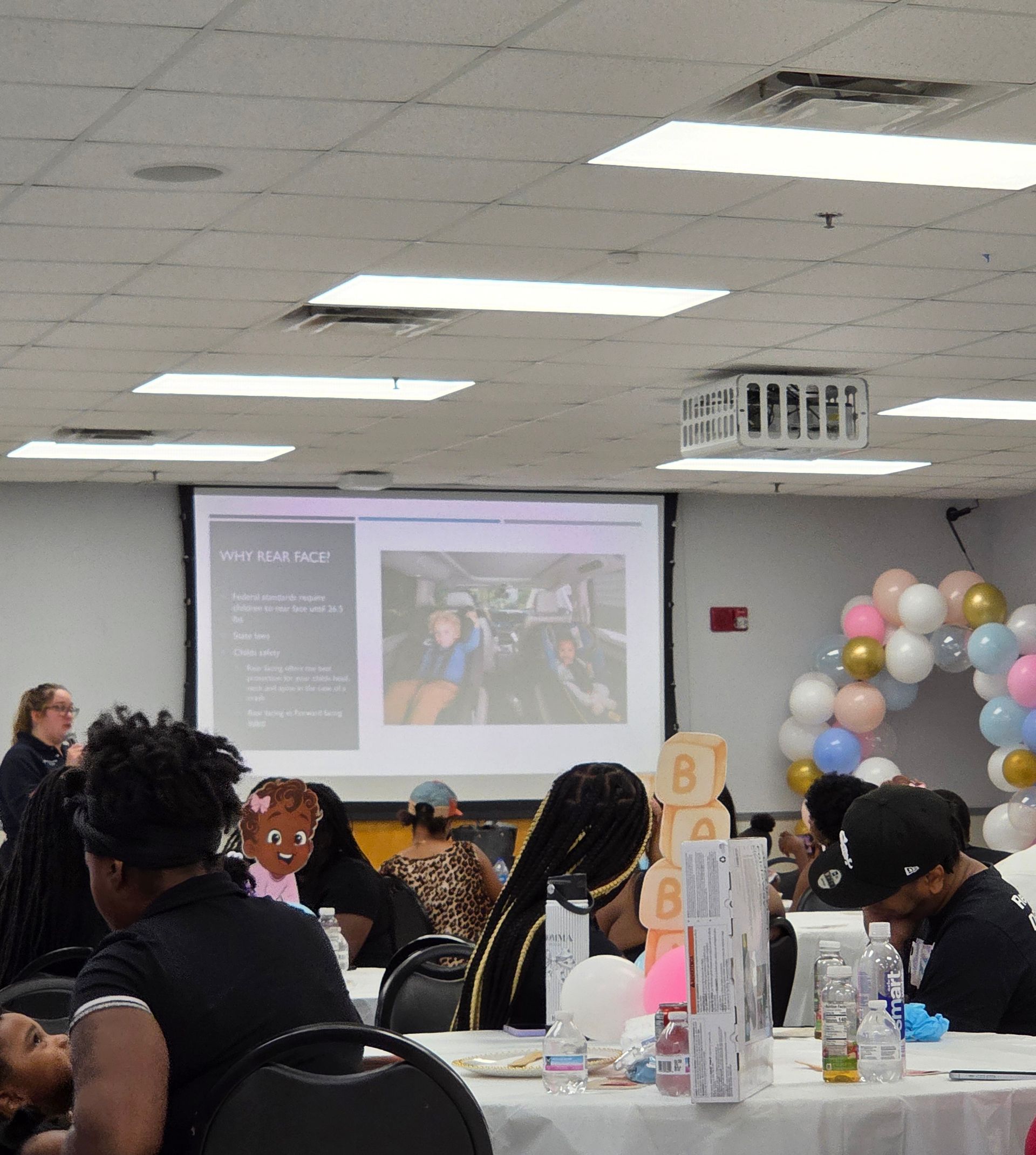 Conference room presentation with seated audience, projected slides, and pastel balloon decorations.
