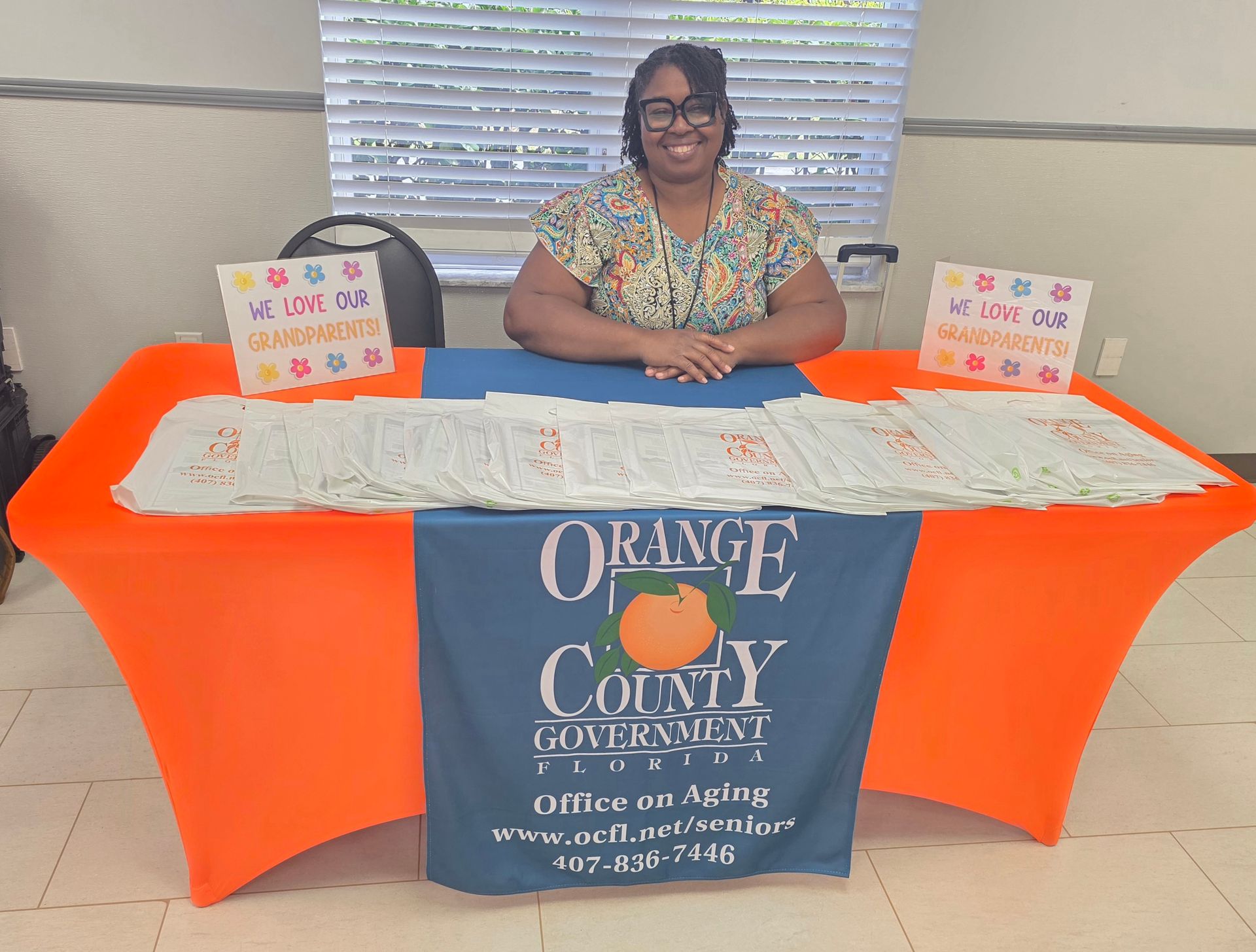 Woman seated behind Orange County government booth with bright orange tablecloth and promotional signs at an indoor event.