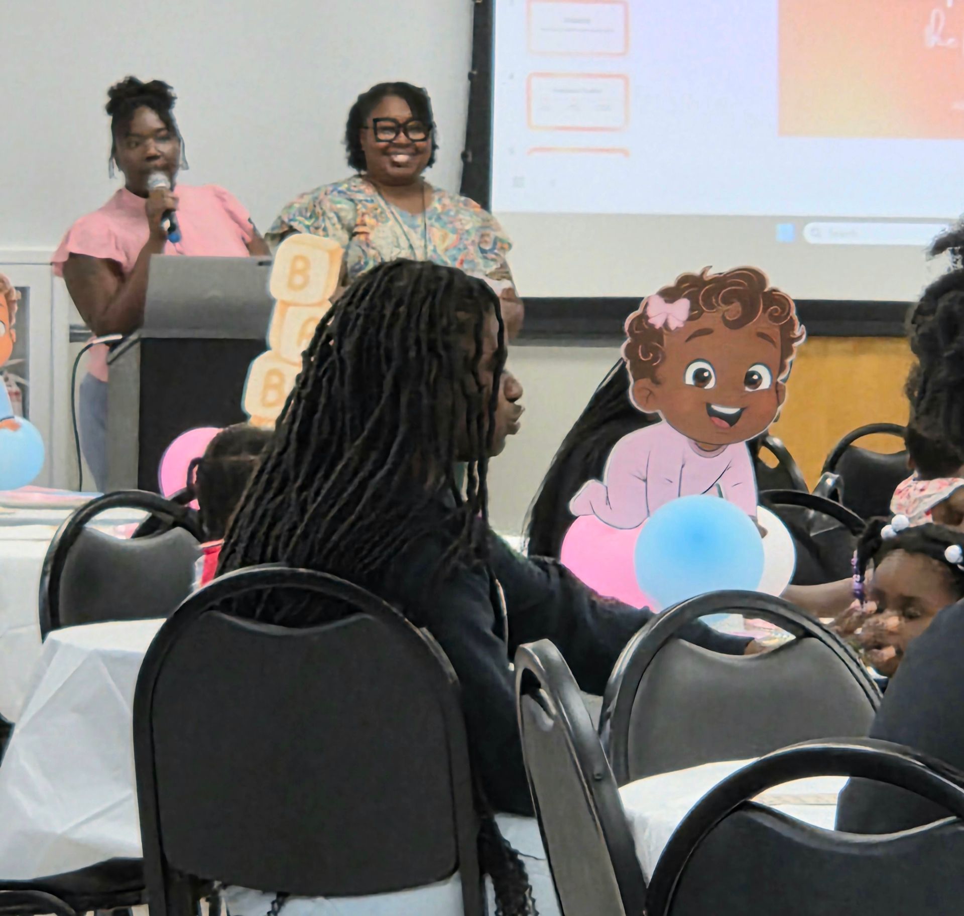 Speakers at a podium in a decorated event room with seated guests and a cartoon child cutout.