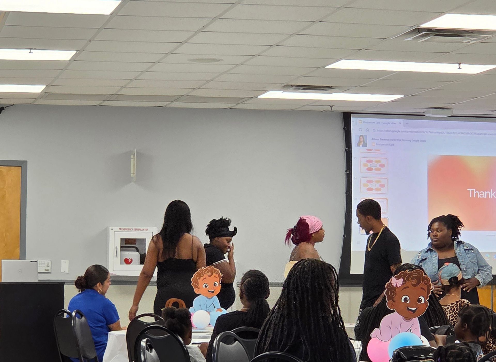 People standing in a classroom or meeting room, facing a projected screen with cartoon cutouts in front.