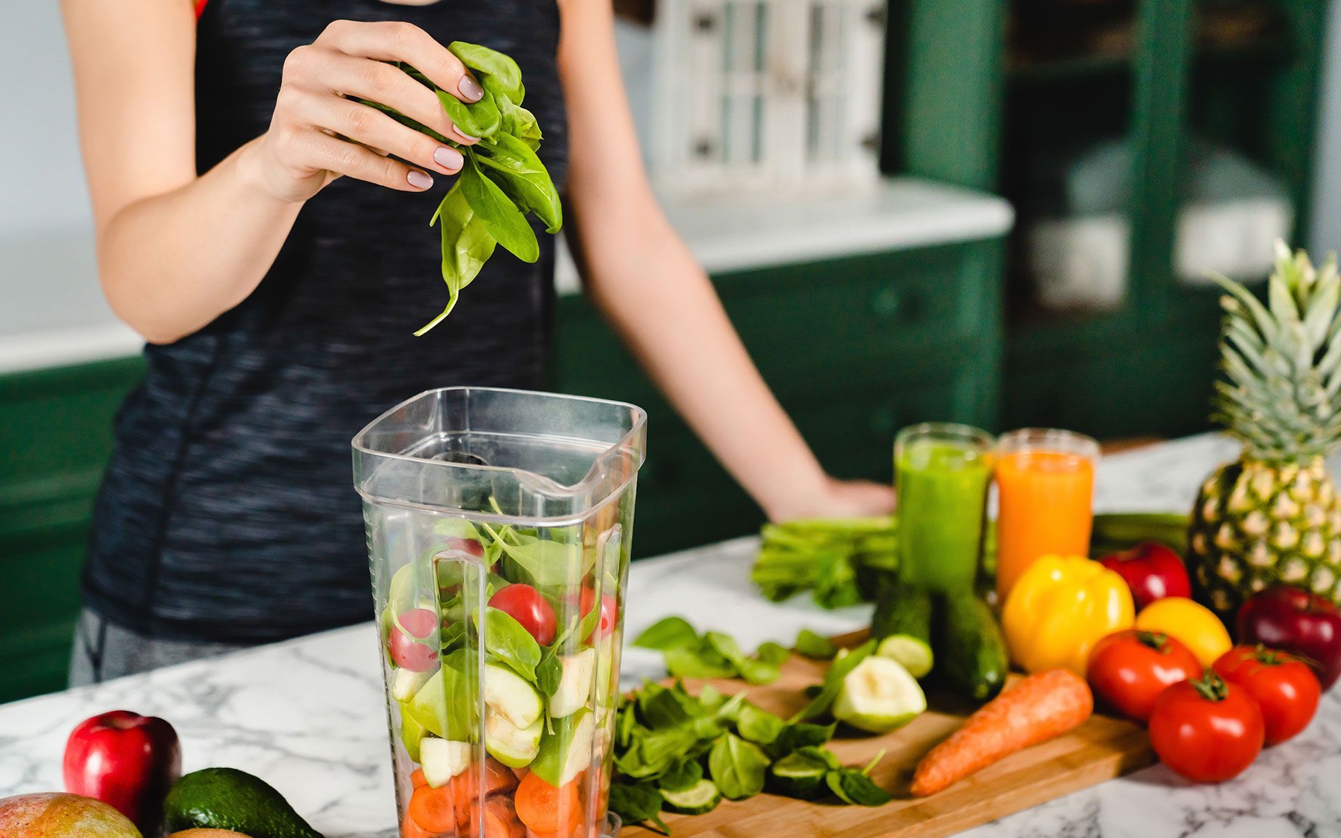 Woman adding spinach to a blender filled with fruits and vegetables. Juices and pineapple nearby.