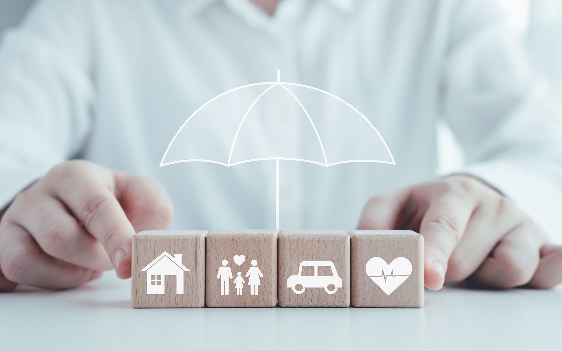 Person holding blocks with illustrations of a house, family, car, and heart beneath an umbrella graphic.
