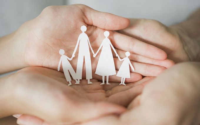 Hands cradling a paper cutout family: parents and two children holding hands.