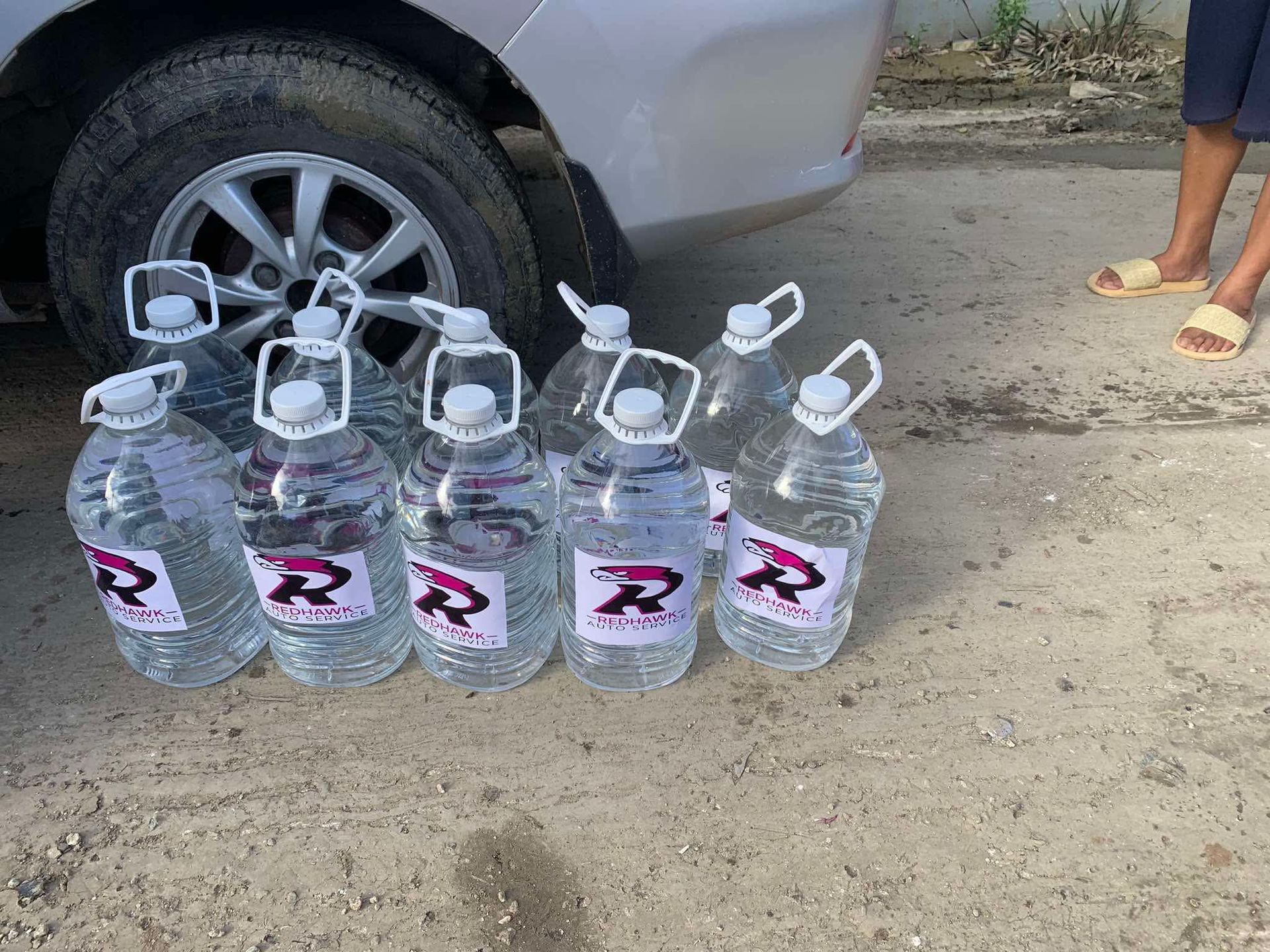 Five clear water jugs lined on a dirt road beside a car tire, with a person's feet visible nearby. | Redhawk Auto Service - Temecula