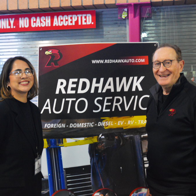 Two people beside a Redhawk Auto Service sign in a garage, both smiling. | Redhawk Auto Service - Temecula
