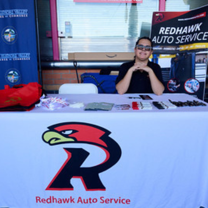 Vendor table with Redhawk Auto Service banner and a person seated behind it at an event booth | Redhawk Auto Service - Temecula
