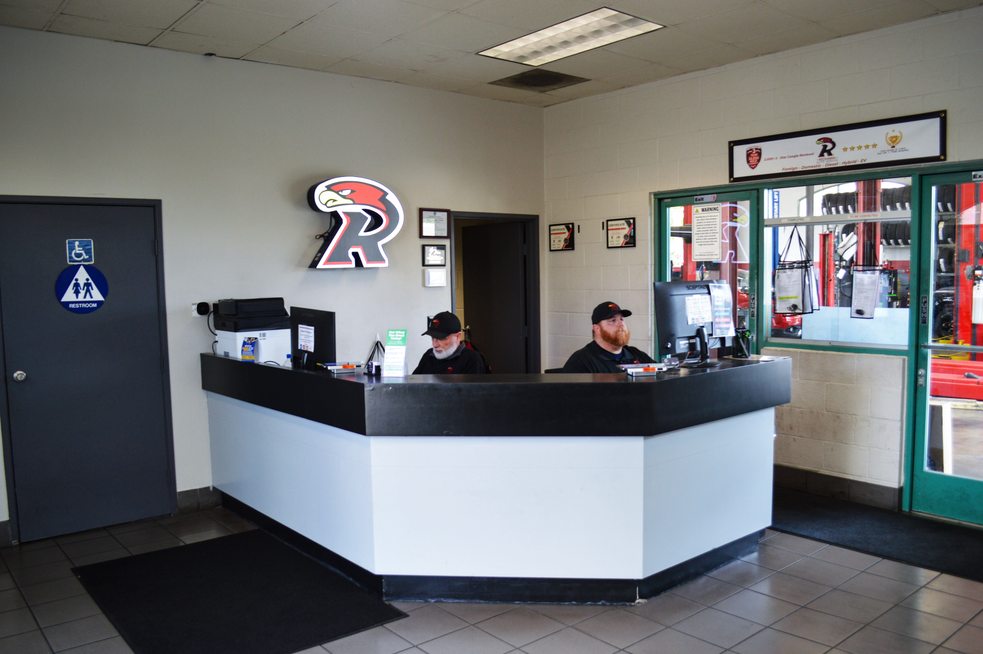 Indoor office lobby with a curved reception desk, two seated staff, and glass entry doors. | Redhawk Auto Service - Temecula