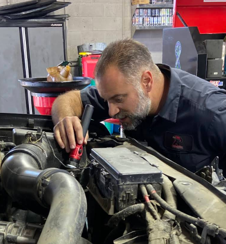 Mechanic inspecting a car engine with a flashlight in a garage, surrounded by tools and equipment | Redhawk Auto Service - Temecula