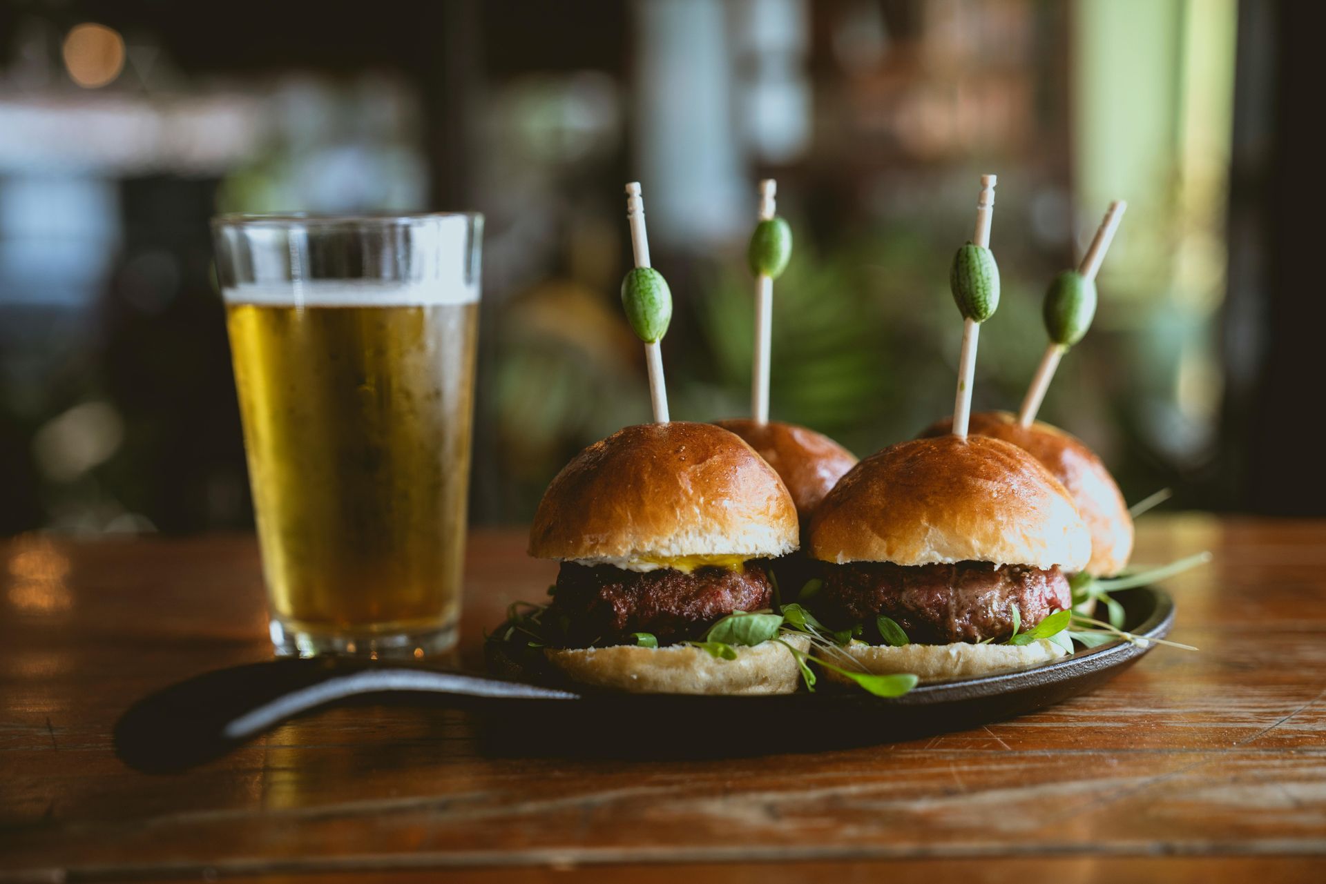 Three sliders on a plate next to a glass of beer on a wooden table.