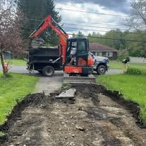 Orange mini excavator loading soil into a black dump truck beside a torn-up driveway in a residential yard