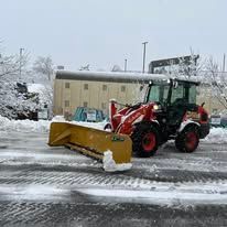 Red tractor with yellow snowplow clearing a snowy parking lot in front of a building.