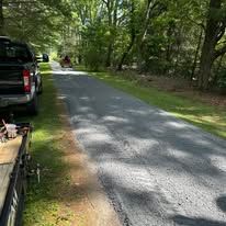 Gravel road in a wooded campground beside parked vehicles and a picnic table