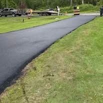 Freshly paved black road curving through a grassy residential area with parked cars in the distance
