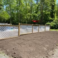 White lattice fence beside a freshly graded dirt yard, with trees and a red wheelbarrow in the background