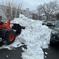 Orange snowplow pushing a large snow pile in a snowy parking lot beside parked cars and leafless trees