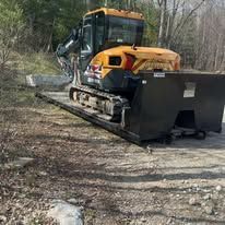 Orange excavator with a front blade clearing a dirt path in a wooded area