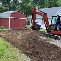Excavator leveling dirt near a red shed and white house in a yard