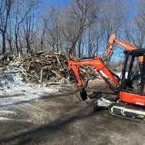 Orange excavator clearing debris beside a snowy roadside and woodpile in a winter wooded area