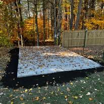 Concrete slab in a wooded backyard, bordered by black tarp and a wooden fence, with autumn leaves scattered around.