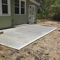 Concrete patio beside a light green house with windows and a door, bordered by dry grass and trees