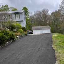 Driveway leading to a white garage beside a house, bordered by trees and a grassy slope.