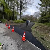 Road repair with fresh black asphalt and orange cones on a wooded driveway, worker in the distance