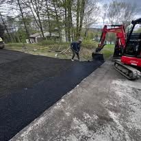 Worker beside red excavator laying black asphalt on a driveway beside a gravel path