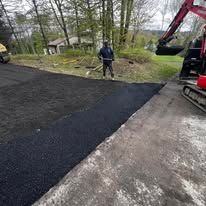 Worker standing on freshly paved driveway beside construction equipment and trees