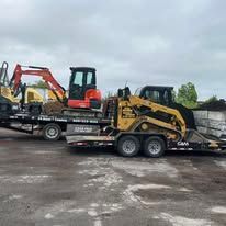 Two compact excavators loaded on a flatbed trailer in a muddy lot under cloudy skies