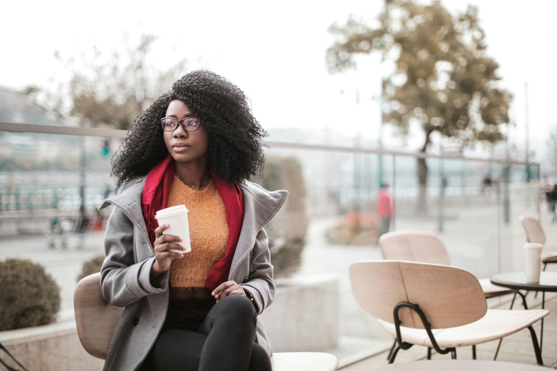 Woman in coat, holding coffee, sits outdoors, looking thoughtful.