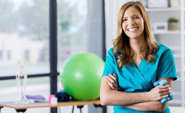 Smiling woman in blue shirt standing with arms crossed in an office or gym setting, with a green exercise ball behind her