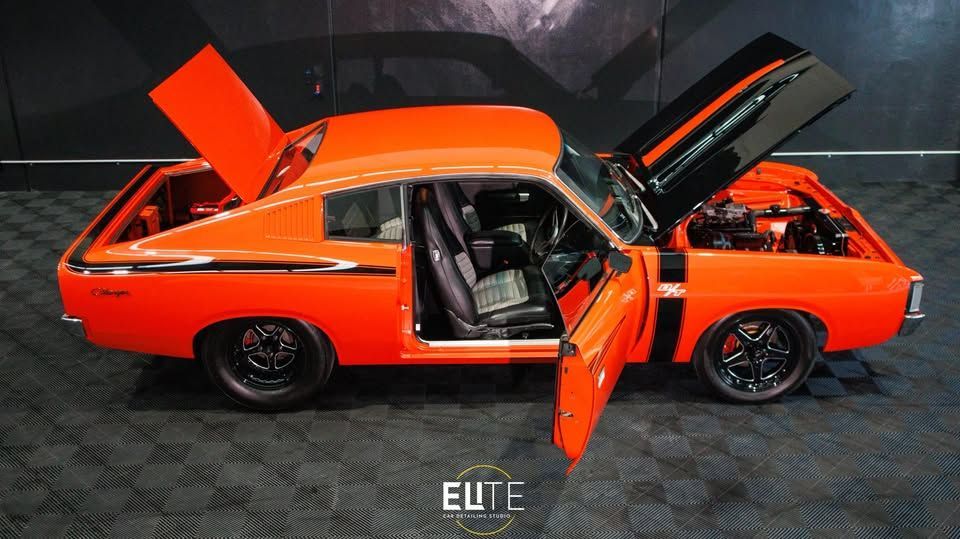 Orange muscle car with open doors and hood, parked inside. Black rims, black stripe, and interior.