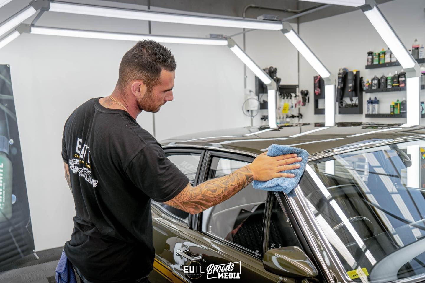 Man in a black shirt wiping a car window with a blue cloth in a well-lit garage.
