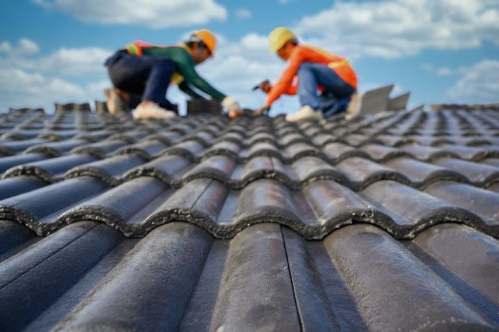 Two workers in safety gear repairing a tiled roof under a blue sky
