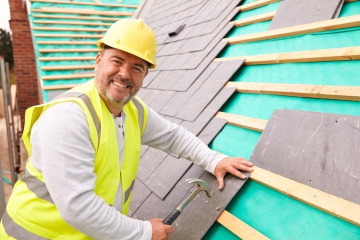 Roofing worker in yellow hard hat installing gray shingles on a teal roof, smiling at the camera