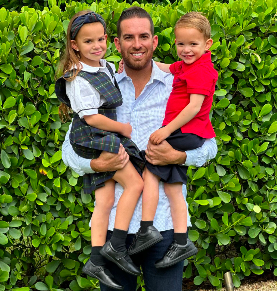A man is holding two children in his arms in front of a hedge.