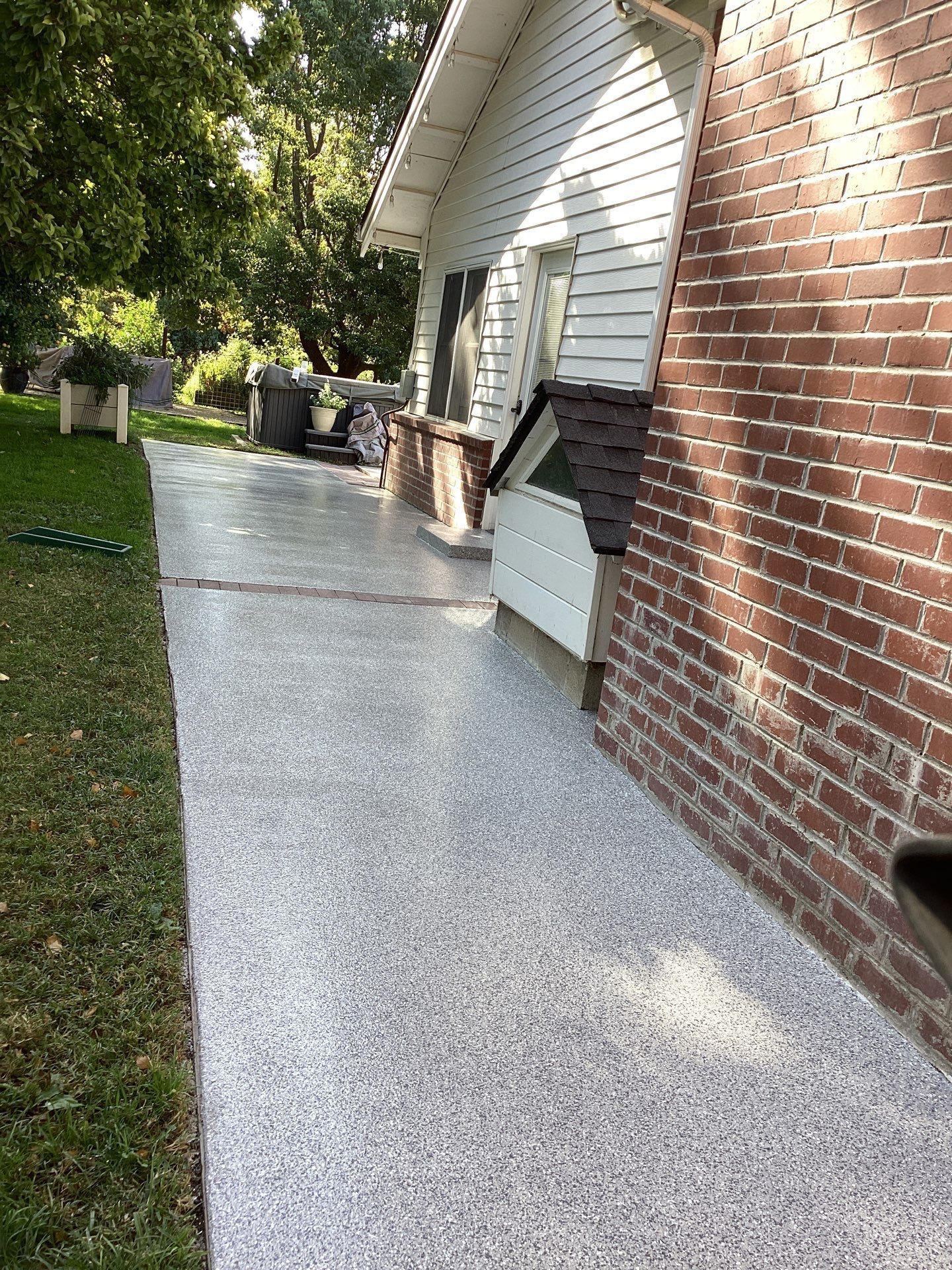 Sidewalk beside a brick house with a white-speckled concrete path and trash bins in the distance