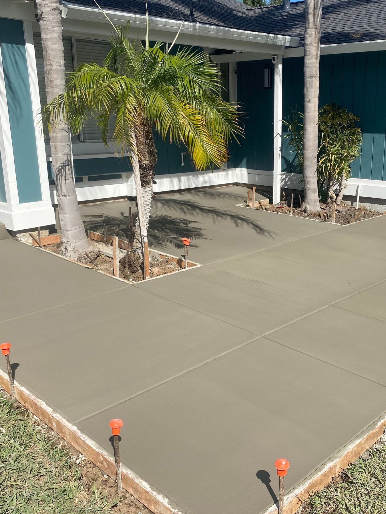 Freshly poured concrete walkway and patio beside a blue house with palm trees and small orange markers