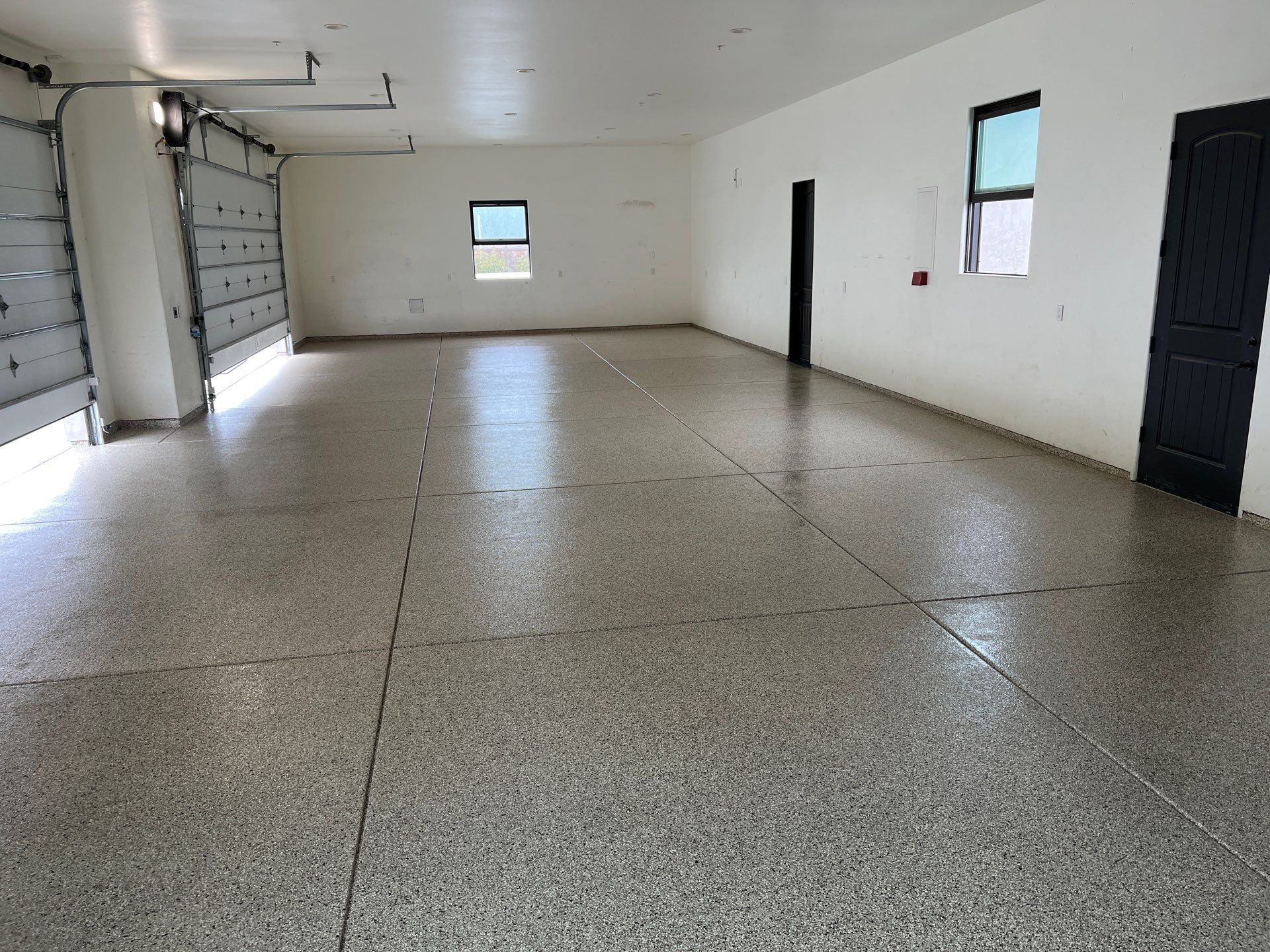 Empty white garage with speckled epoxy floor, a dark door, small windows, and string lights along the left wall