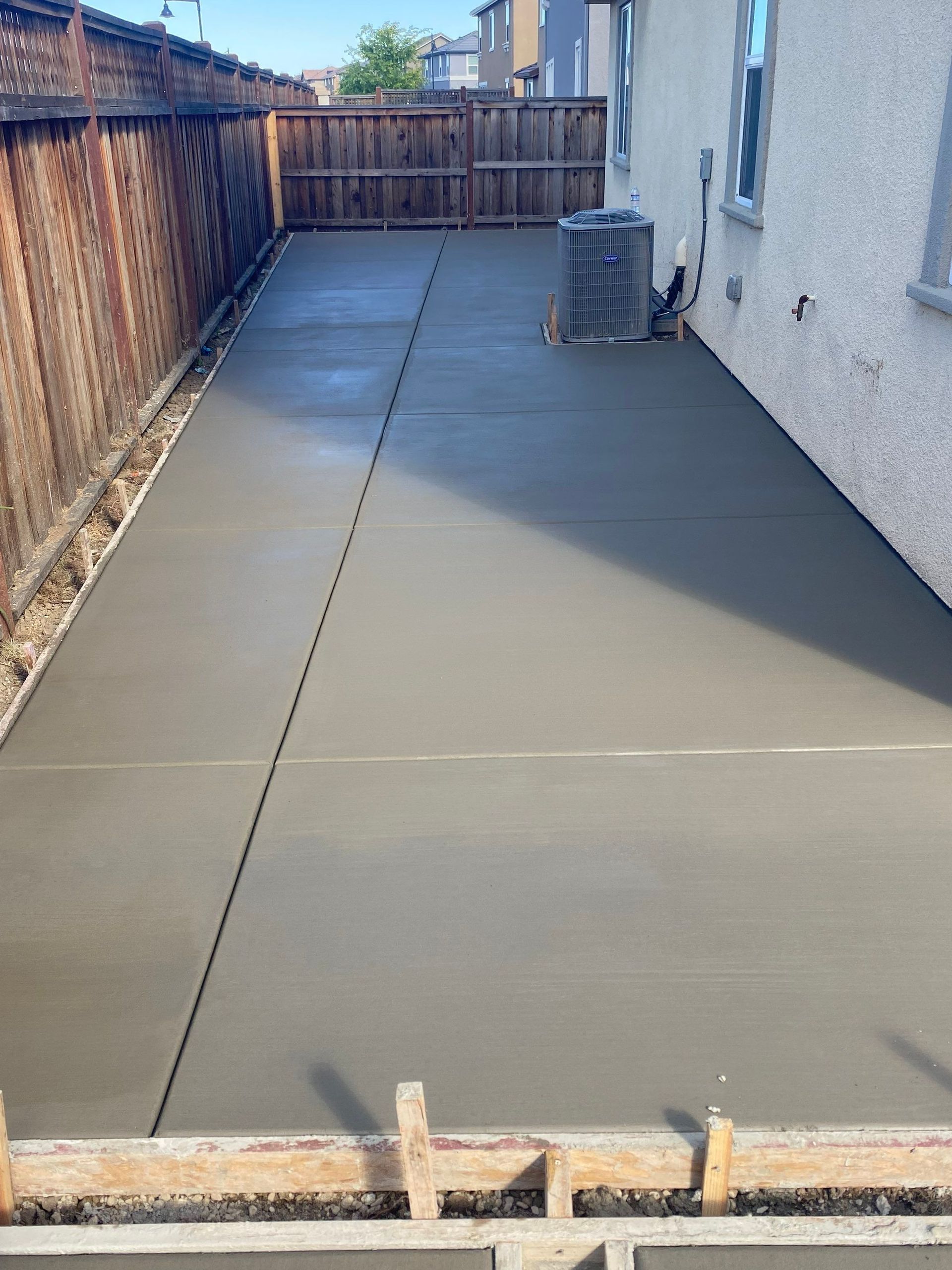 Narrow concrete backyard walkway with wooden fence, house wall, and air conditioner unit at the far end