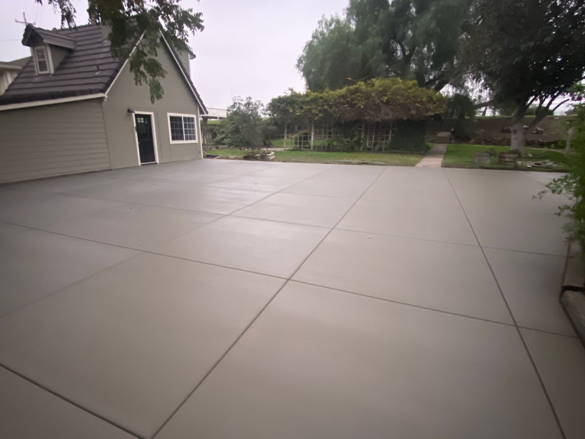 Wide concrete driveway beside a beige house with trees and a garden in the background