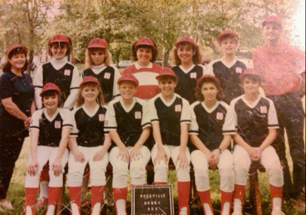 Youth baseball team posing in uniforms with coaches, holding a trophy outdoors.