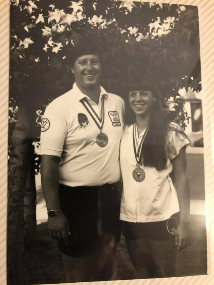Two medal winners posing outdoors, smiling in matching white shirts and black hats.