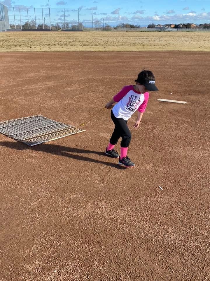 Child in a pink shirt running on a dirt baseball field near a base, with open sky in the background
