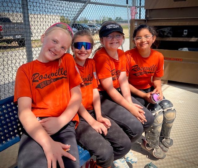 Four softball players in orange jerseys sit on a bench, smiling in the sun at a field.