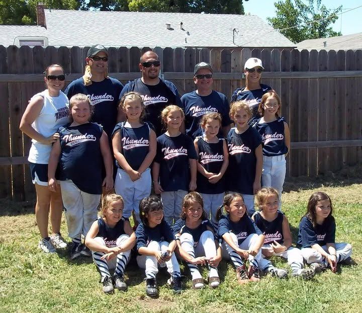 Youth softball team and coaches posing together outdoors in matching navy shirts in front of a wooden fence.
