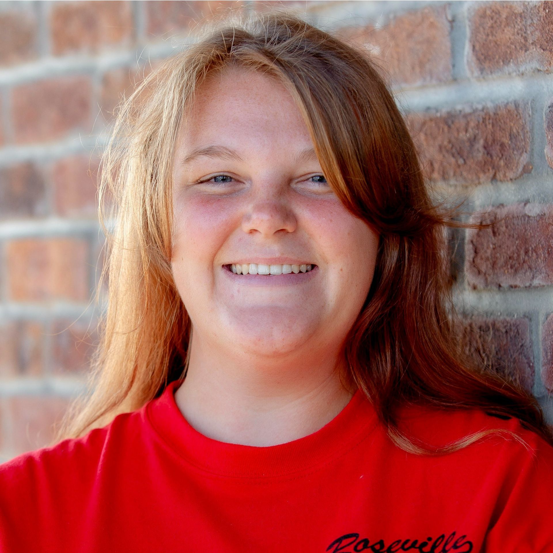 Smiling person in a red shirt standing in front of a brick wall