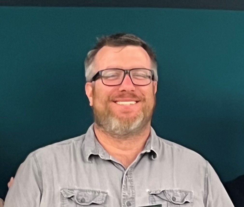Smiling man with glasses and beard, wearing a gray shirt against a teal background