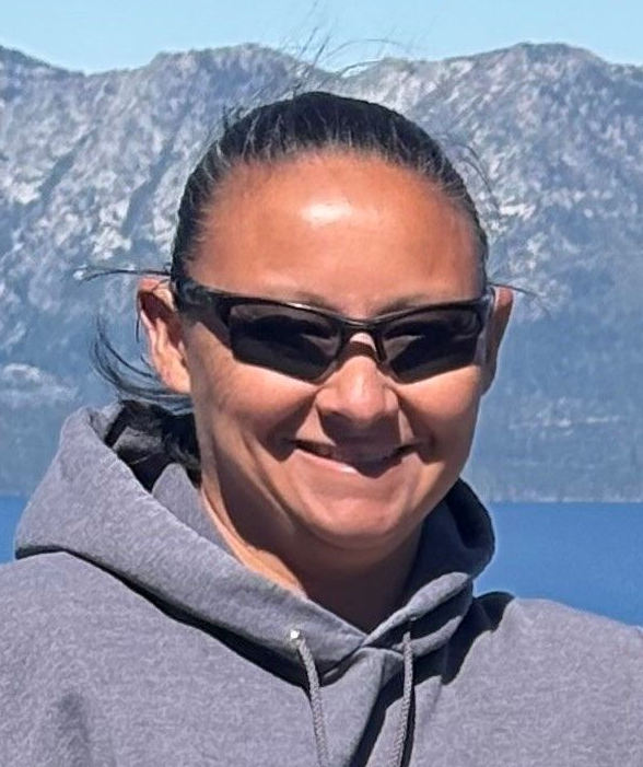Smiling person in sunglasses and gray hoodie with mountains and blue water in the background